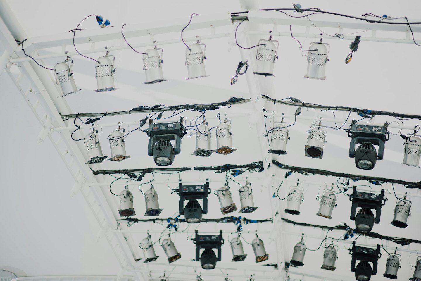 Stage lights hang from a white truss with many black and blue wires.